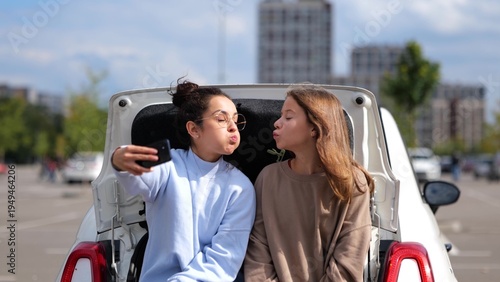 Beautiful happy teen girl sitting in car trunk with pretty mom taking selfie photos on smartphone. Caucasian woman with daughter or sister making funny face expression posing to cellphone