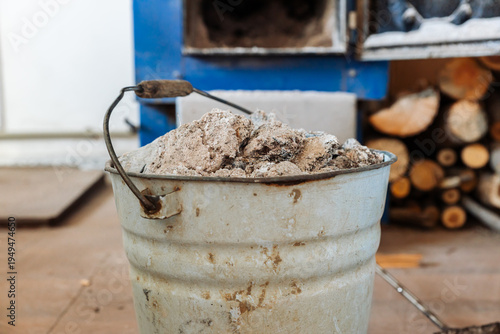 A metal ash bucket stands in front of a blue wood-burning stove with an open door. A stack of chopped firewood lies on the floor.