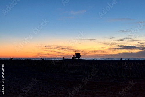 Silhouette of a lifeguard tower at Santa Monica Beach after sunset, glowing evening sky and warm holiday atmosphere over the calm Pacific coast.
