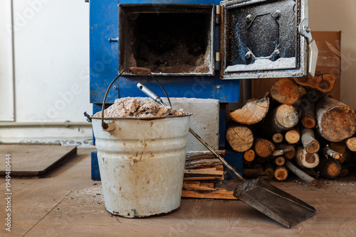 A blue wood-burning stove with an open door. A stack of chopped firewood lies on the floor. A shovel is next to a metal bucket of ashes.