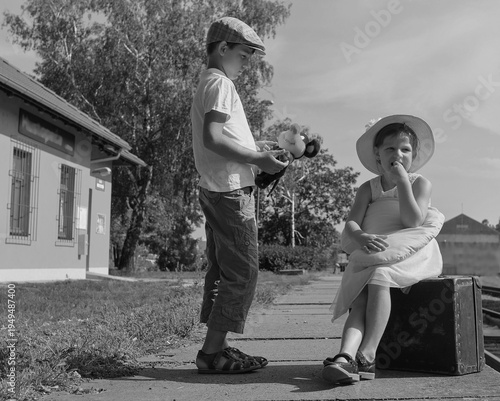 Brother and sister waiting at a train station with luggage and toys, vintage style