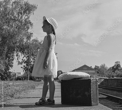 Young girl in a dress and hat waits with a vintage suitcase on a train platform