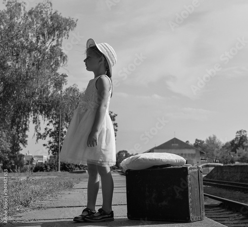 A sad little girl in a white dress with a stuffed toy and an old suitcase. The adorable sad young girl stands on a train station platform. The concept of the end of summer vacation