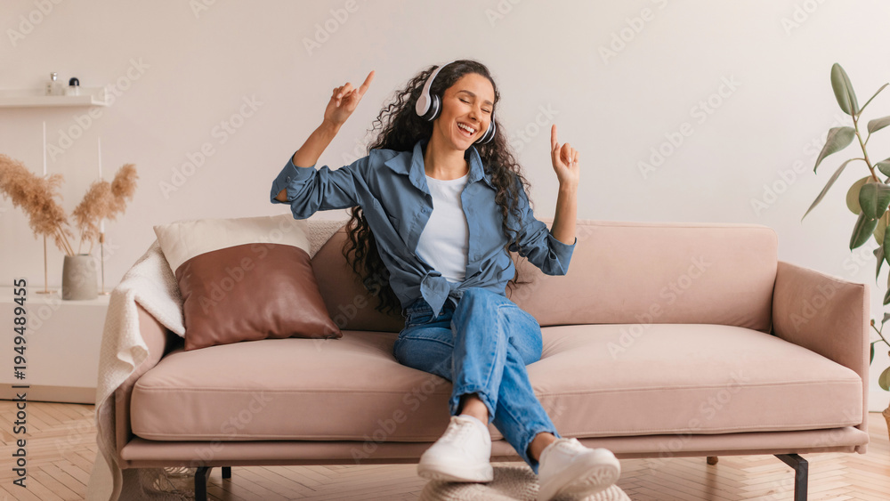 Obraz premium A woman sits on a sofa in her living room wearing headphones. She smiles and dances to music, enjoying her time at home. Light colors fill the space, enhancing the mood of relaxation.