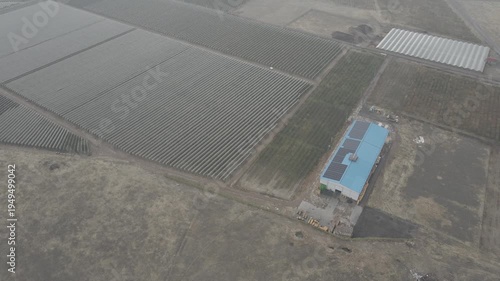 Aerial drone footage over extensive agricultural fields covered with white netting, approaching a large industrial farm building with solar panels on its blue roof under an overcast sky.

