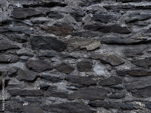 Dark natural stone wall texture with layered slate rocks and rough mortar, creating a rugged rustic background ideal for architectural and design use.