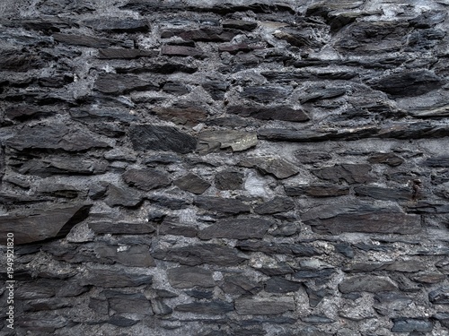 Dark slate stone wall texture with layered natural rock pieces and rough mortar, creating a dramatic rustic background for design, architecture, or construction themes.