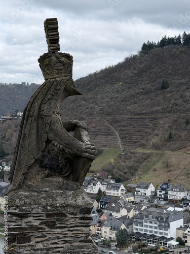 Weathered stone gargoyle overlooking a hillside town under dramatic cloudy skies, blending medieval architectural sculpture with a moody European landscape.