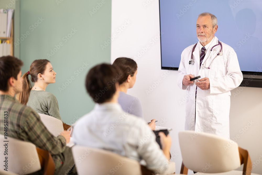 Fototapeta premium Elderly male medical professor in medical coat gives lecture to group of male and female students in university auditorium