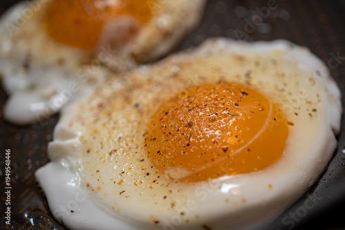 Two sunny side up fried eggs in a pan seasoned with black pepper and spices. Close-up of fresh organic breakfast cooking. Healthy protein meal background
