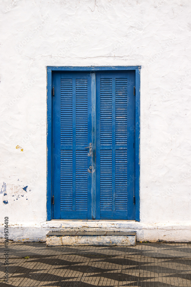Fototapeta premium A weathered blue louvered double door set in a textured white plaster wall with a small concrete step and patterned tiled pavement in front.