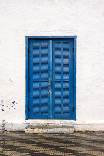 Bild auf Leinwand A weathered blue louvered double door set in a textured white plaster wall with a small concrete step and patterned tiled pavement in front