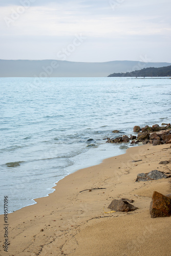 Canvas Print Sandy shoreline with scattered rocks and gentle waves lapping a calm blue sea under a pale sky with distant hills visible on the horizon