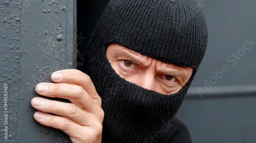 A suspicious man wearing a black balaclava peeks around a corner while hiding from someone.