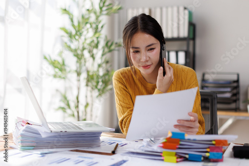 Fotografi A focused woman wearing a yellow sweater reviews documents while on a phone call in a bright office space filled with organized paperwork and a laptop