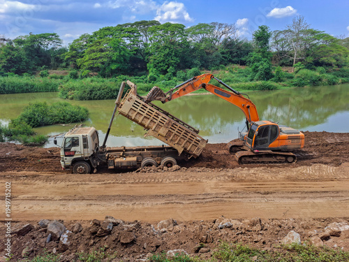 Construction work is taking place by the river; trucks are lifting their trailers to unload soil, and an orange backhoe is parked nearby, assisting in clearing the earth.
