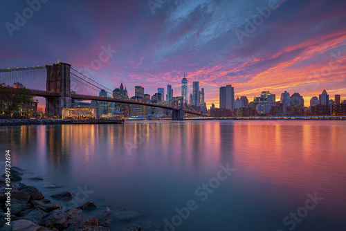 Sunset over wide waterfront panorama of city skyline with large suspension bridge and colorful sky reflecting on calm water, creating peaceful urban scene