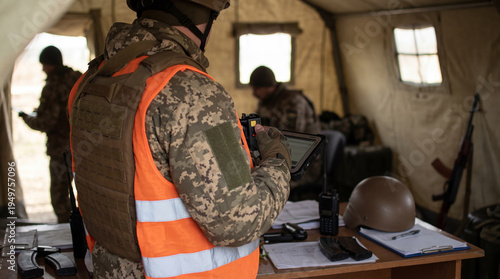 Photos Military officer in camouflage uniform and orange vest using a digital tablet inside a tactical command tent