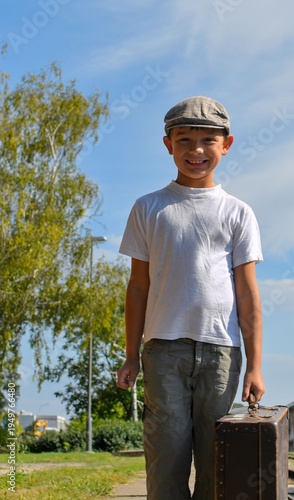 A cute boy at the train station. A little smiling boy holds an old suitcase. The concept of travel, vacation, and carefree childhood. Retro and vintage vibe.