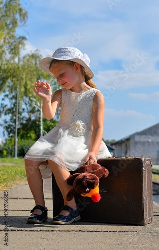 A cute small girl in a white dress with a plush toy and an old suitcase. The adorable smiling girl is standing on a train platform. The concept of the end of summer vacation, traveling, and a carefree