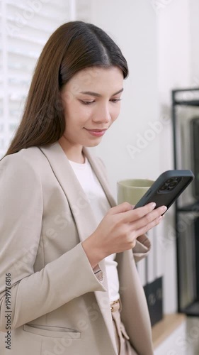 Asian businesswoman smiling while using smartphone holding coffee cup near window, adult female professional checking messages during short break, communication, digital connection relaxation at work