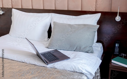 laptop working on white bed, pillows in modern hotel bedroom, comfortable workspace setup on bedding with soft textile blanket wooden headboard, remote work, hospitality business, travel