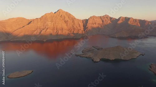 Drone flying over calm waters of the Strait of Hormuz between Oman and Iran near Musandam Peninsula with dramatic desert mountains at sunset, a strategic global oil shipping route.