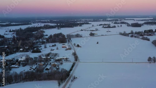Winter expanse covering houses, fields, and roads during twilight glow in aerial view with snowfall and pink sky