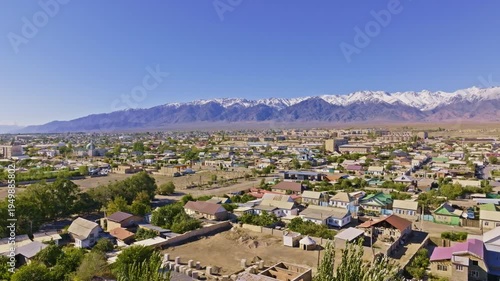 Aerial view captures serene Balykchy town surrounded by agricultural land and trees. Snow-capped mountains rise majestically against a clear blue sky.