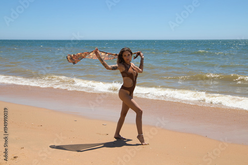 A beautiful young woman with tattoos and a swimsuit walks along the shore with a sarong fluttering in the wind. The woman enjoys the solitude and tranquility of the sunny day.