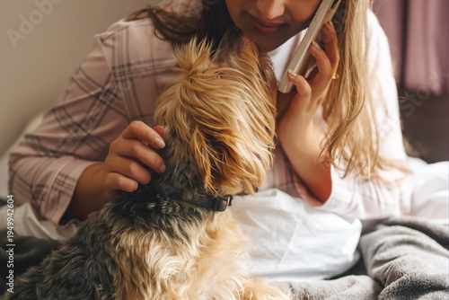 Woman talks on phone while petting a small dog on a bed in a cozy room during daytime