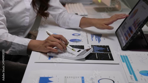 Financial analyst calculating business data and reviewing graphs on a tablet and computer while working overtime in office.