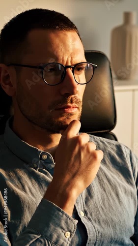 A pensive man thinking hard sits in an office chair. The serious man touches his chin while thinking about a business problem. A pensive look crosses the face of the thinking man. He looks focused.