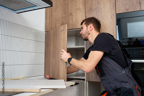 Foto Man in overalls installing wooden kitchen cabinet door in modern apartment interior