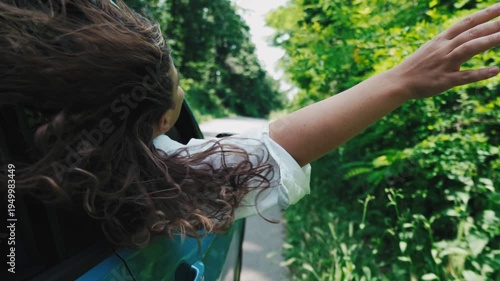 Young curly-haired woman leaning out of a car window, hair blowing in the wind, enjoying a scenic road trip through nature. Freedom, travel lifestyle and carefree summer vibes.