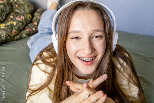 Portrait of a delighted teenage girl with braces wearing headphones lying on bed at home. Relaxed student lifestyle with technology, music listening, online learning and modern digital youth culture.