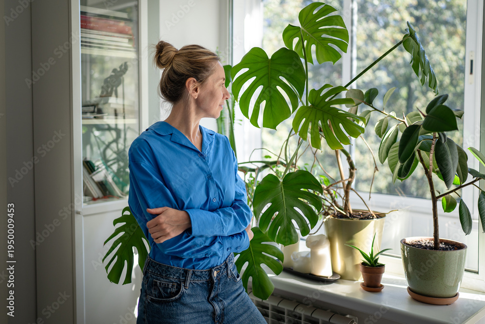 Obraz premium Thoughtful positive middle aged woman florist looking at own indoor garden on windowsill, observing plants with love and care. Mature female plant lover posing surrounded by potted houseplants at home