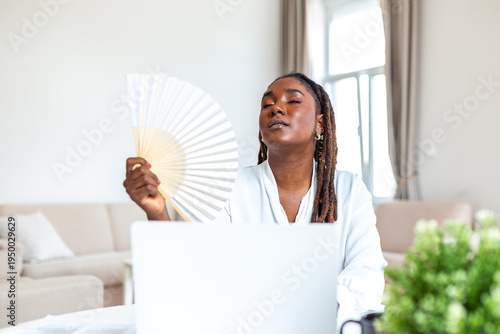 Φωτογραφία Overheated black woman using hand fan while working at laptop in summer