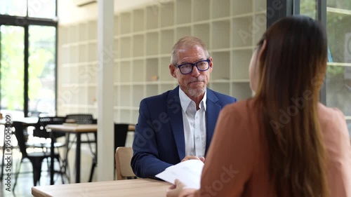 Experienced male hiring manager in a suit asking questions and listening to a young woman's answers during a job interview in a modern office space, focusing on her curriculum vitae