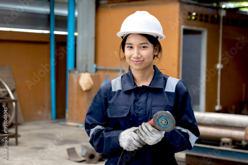 Portrait of happy Asian industrial female engineer supervisor in safety suit and white helmet holds metal polishing machine at manufacturing plant. Cheerful female worker working at metalwork factory.