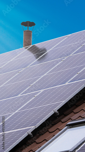 Solar panels collect sunlight on a roof in a clear sky during the day near a chimney