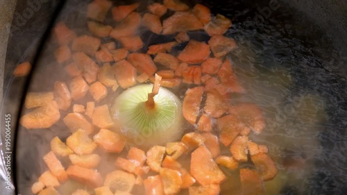 Top view close-up of chopped carrots and a whole onion boiling in a cauldron with hot steam.