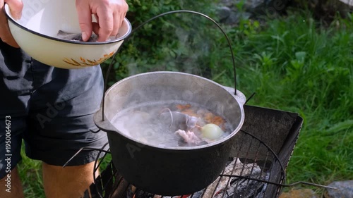 Man adding fresh fish into boiling cauldron with vegetables over open fire outdoors, medium shot.
