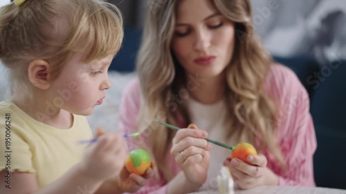 Mother and child paint colorful eggs together. Soft light shows calm, joyful moments. Child holds egg, concentrates. Mother guides, smiles gently. Perfect for Easter, family bonding, creativity themes