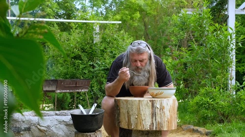 Senior bearded man eating fresh fish soup from a clay bowl sitting at a wooden stump table in a summer garden.