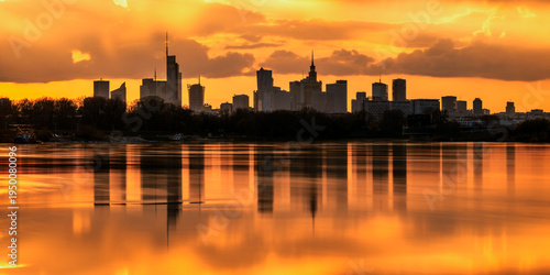 View of the city skyline mirrored in the tranquil water under a fiery sunset glow, silhouetting buildings against the bright sky, Warsaw, Masovian Voivodeship, Poland.