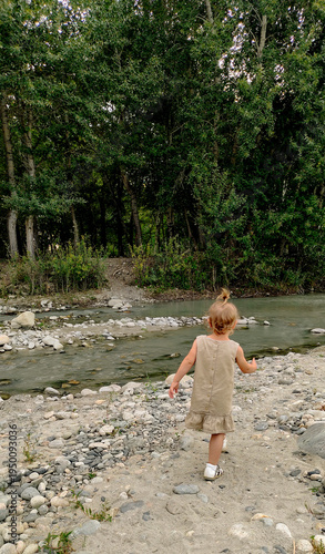 A little girl plays on the bank of a mountain river on a cloudy evening. She runs and stands by the water. An atmosphere of calm, natural freedom, the joy of childhood, and closeness to nature.