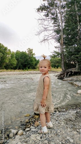 A little girl plays on the bank of a mountain river on a cloudy evening. She runs and stands by the water. An atmosphere of calm, natural freedom, the joy of childhood, and closeness to nature.