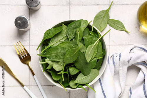 Bowl with fresh spinach leaves and cutlery on white tile background