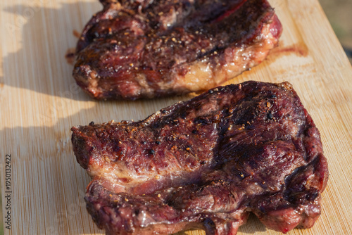 two seasoned, freshly grilled steaks resting on a light wooden board, highlighting the charred exterior and peppercorn seasoning under bright natural sunlight.